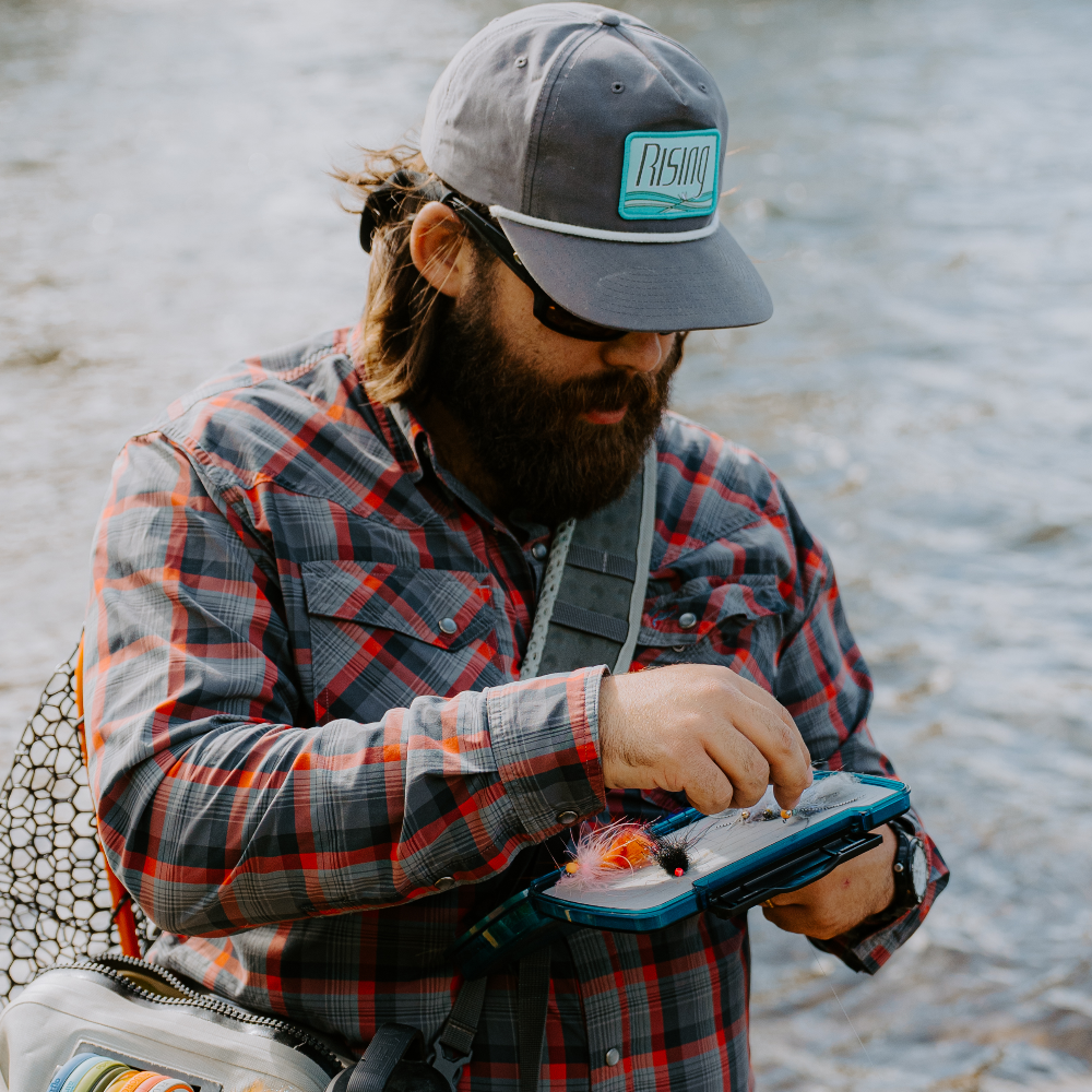 Man fishing by a river, wearing a cap and plaid jacket.